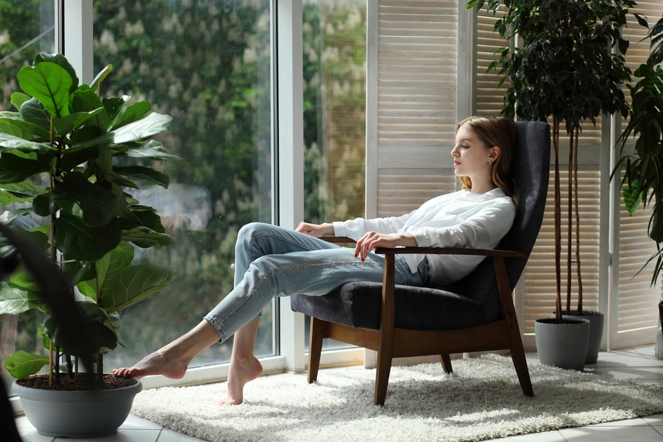 Femme détendue assise dans un fauteuil moderne près d'une baie vitrée, entourée de plantes vertes d'intérieur