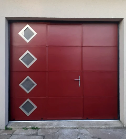 Porte de garage sectionnelle rouge bordeaux avec quatre hublots losangés en verre fumé encadrés de blanc, équipée d'une poignée métallique