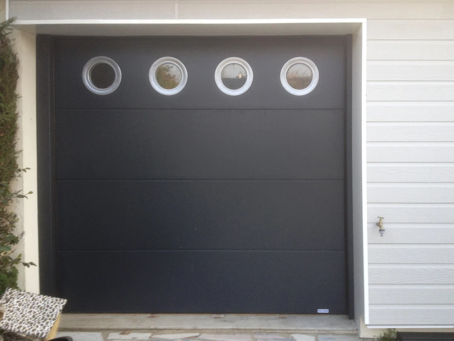 Porte de garage sectionnelle moderne anthracite avec quatre hublots circulaires en verre, installée sur façade de maison contemporaine